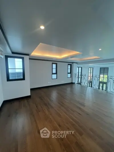 Spacious and elegant living room with polished hardwood floors, sophisticated tray ceiling design, and abundant natural light from multiple windows leading to a charming balcony.