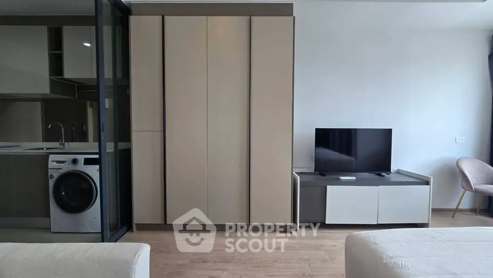 Modern living room with sleek cabinetry, TV unit, and adjacent kitchen featuring a washing machine.