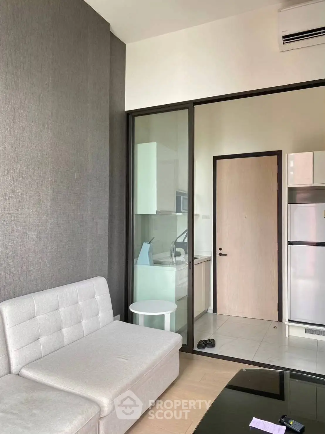 Modern living room with sleek sofa and glass partition leading to kitchen area.