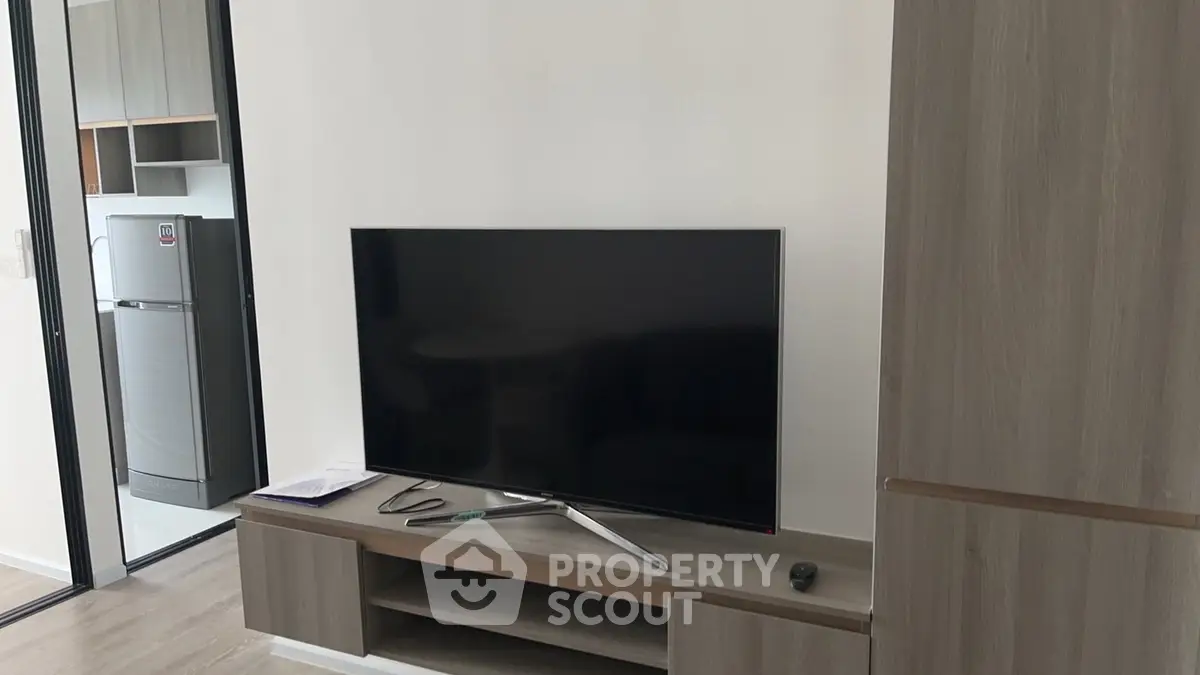 Modern living room with sleek TV stand and large flat-screen TV, adjacent to a kitchen with stainless steel fridge.