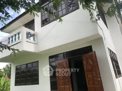 Charming two-story house with wooden doors and balcony, surrounded by lush greenery.