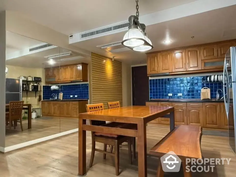 Spacious kitchen with wooden cabinets and dining area, featuring modern lighting and blue backsplash.