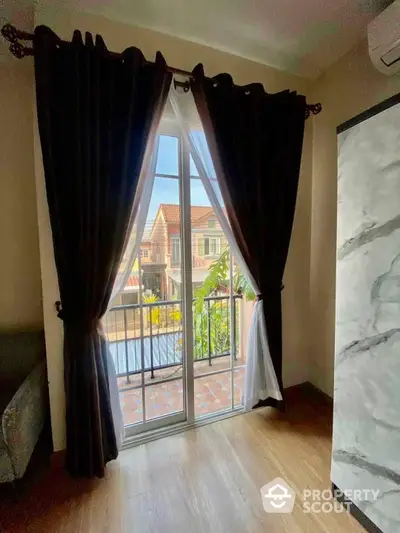 Charming room with elegant dark curtains opening to a sunlit balcony overlooking a serene neighborhood, featuring sleek wooden flooring and a touch of modern marble wall design.