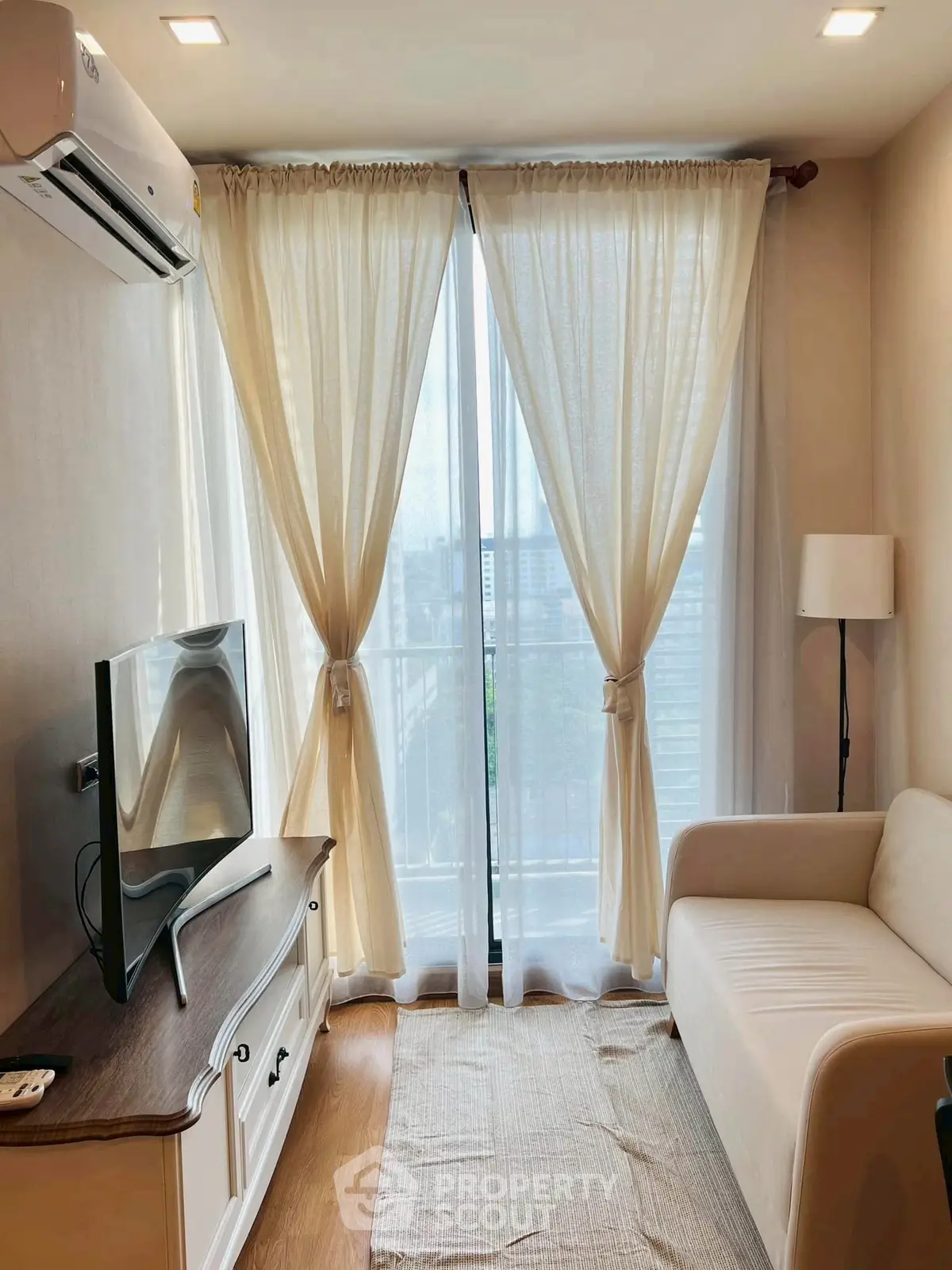 Cozy living room with natural light, featuring a sofa, TV, and elegant curtains leading to a balcony view.