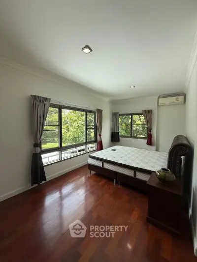 Spacious bedroom with large windows and hardwood floors, offering a serene view of greenery.