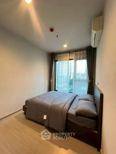 Modern bedroom with large window and city view, featuring cozy bedding and neutral decor.