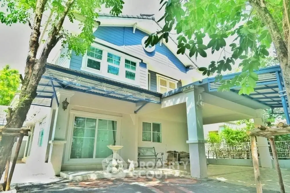 Charming two-story house with blue accents and spacious carport surrounded by lush greenery.