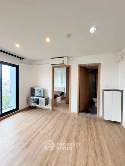 Modern apartment interior with wooden flooring, large windows, and a glimpse of a bedroom and bathroom.