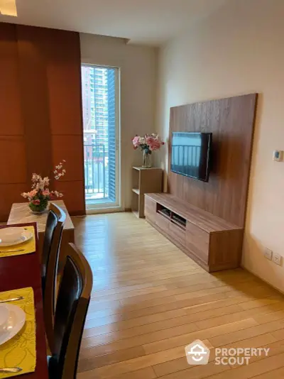 Cozy living room with warm wooden floors and furnishings, leading to a sunlit balcony with urban views, perfect for relaxed city living.