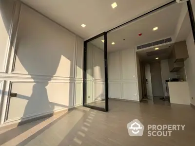 Sunlit spacious living room with large windows and a glass door leading to a balcony, featuring modern flooring and a view into a sleek kitchen area.