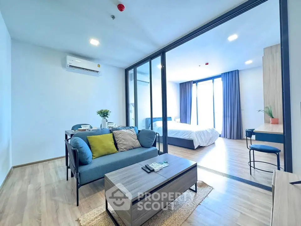 Modern open-plan living room with glass partition leading to a bright bedroom, featuring stylish furniture and decor.