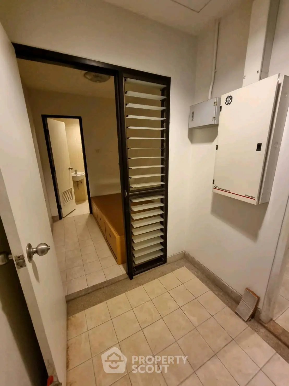 Compact storage room with tiled floor and utility panel, adjacent to a small bathroom.