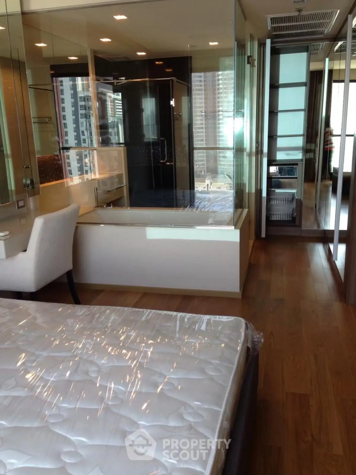 Modern bedroom with glass-walled bathroom and city view, featuring hardwood floors and elegant furnishings.