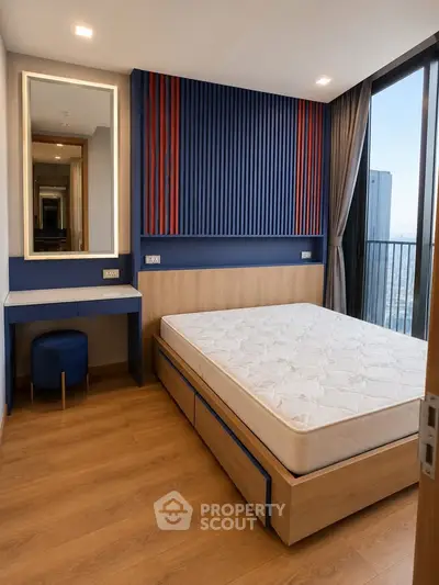 Modern bedroom with stylish decor and large window offering city views, featuring a comfortable bed and vanity area.