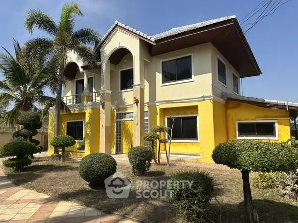 Charming two-story yellow house with manicured garden and palm trees under clear blue sky.
