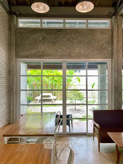 Charming interior with large windows opening to a lush garden view, featuring rustic furniture and industrial design.