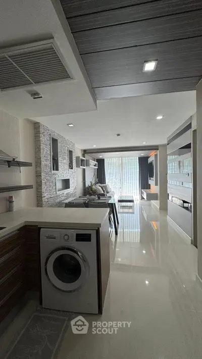 Modern open-plan living room with sleek kitchen and washing machine, featuring stylish decor and ample natural light.