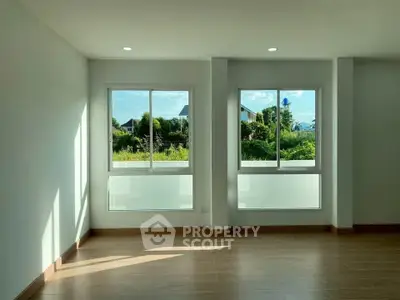 Bright empty room with large windows and wooden flooring, offering a serene view of greenery outside.