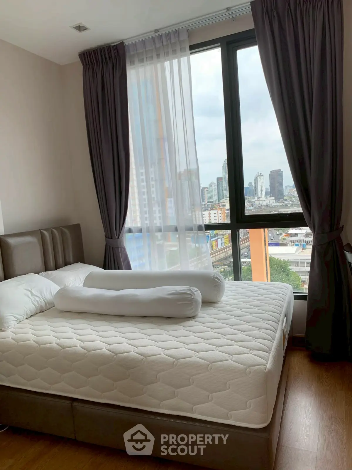 Modern bedroom with large window offering city skyline view, featuring cozy bed and elegant curtains.