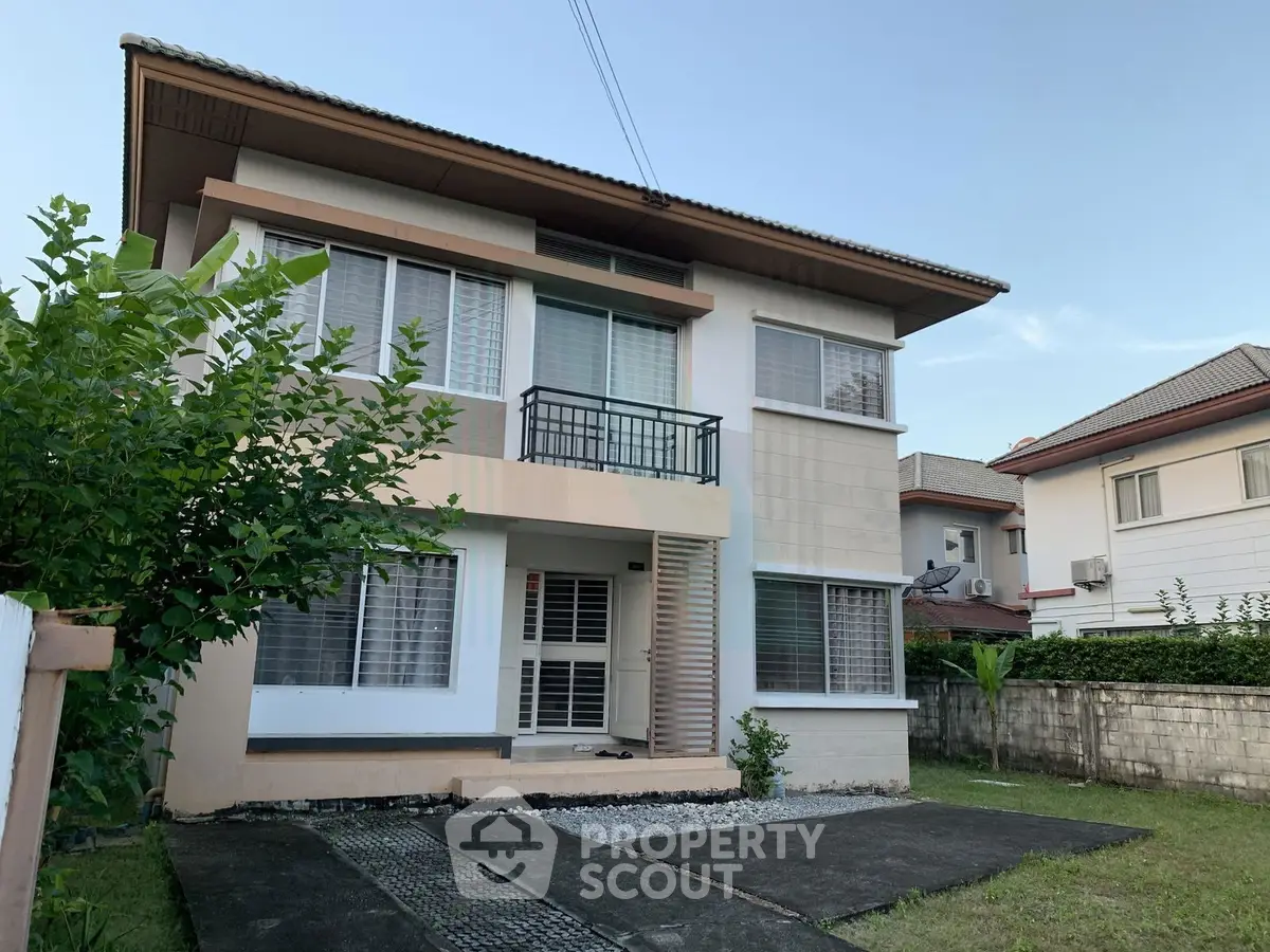 Modern two-story house with large windows and a small balcony, surrounded by greenery.