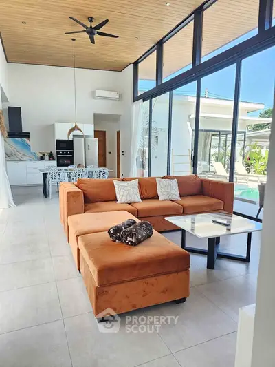 Luxurious open-plan living room with modern kitchen and pool view, featuring stylish orange sofa and high ceilings.