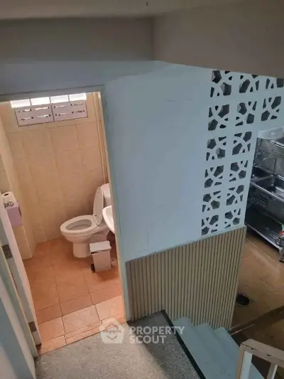 Compact bathroom with tiled walls and toilet, adjacent to a staircase and kitchen area.