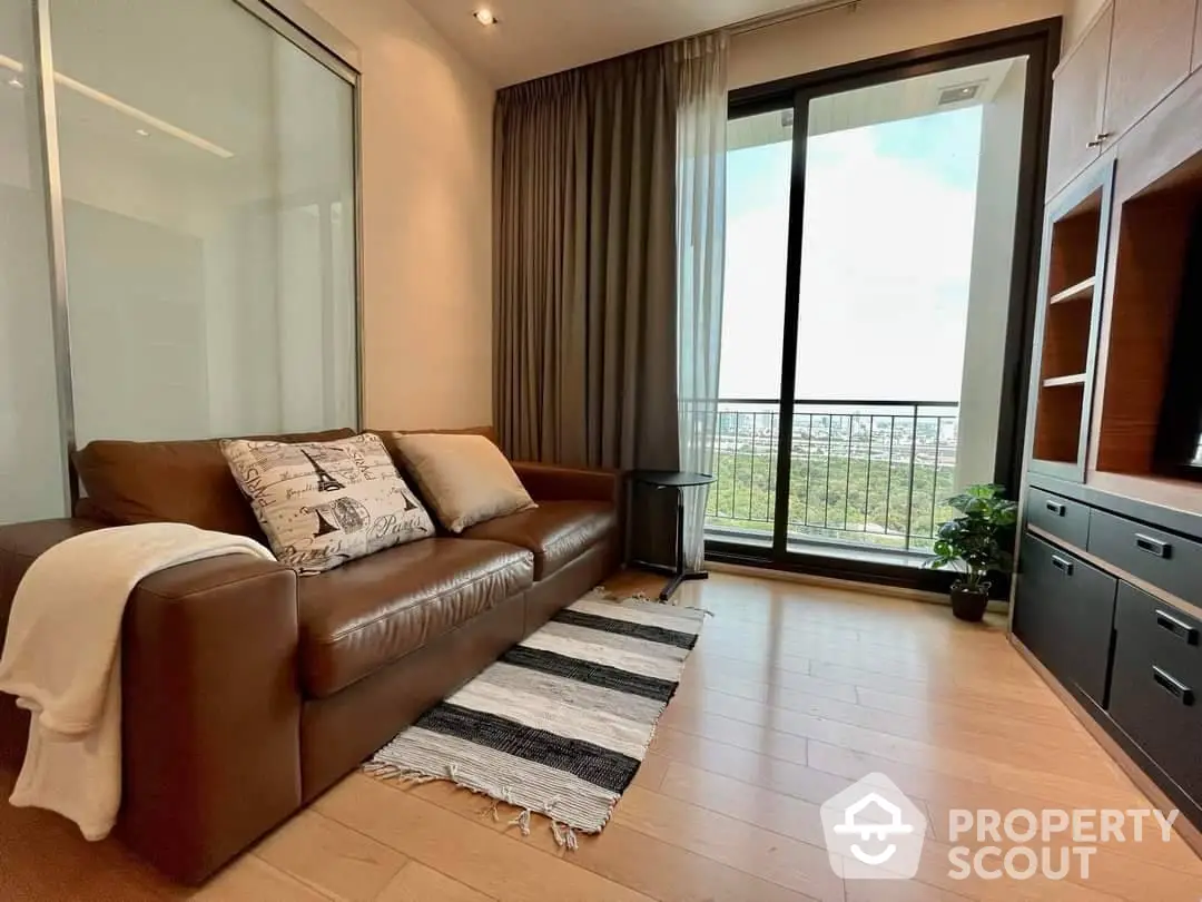 Spacious living room with large windows leading to balcony, offering natural light and a comfortable brown leather sofa, perfect for relaxing.