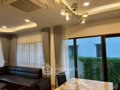 Elegant living room with modern lighting and large windows overlooking greenery, featuring stylish curtains and a marble table.