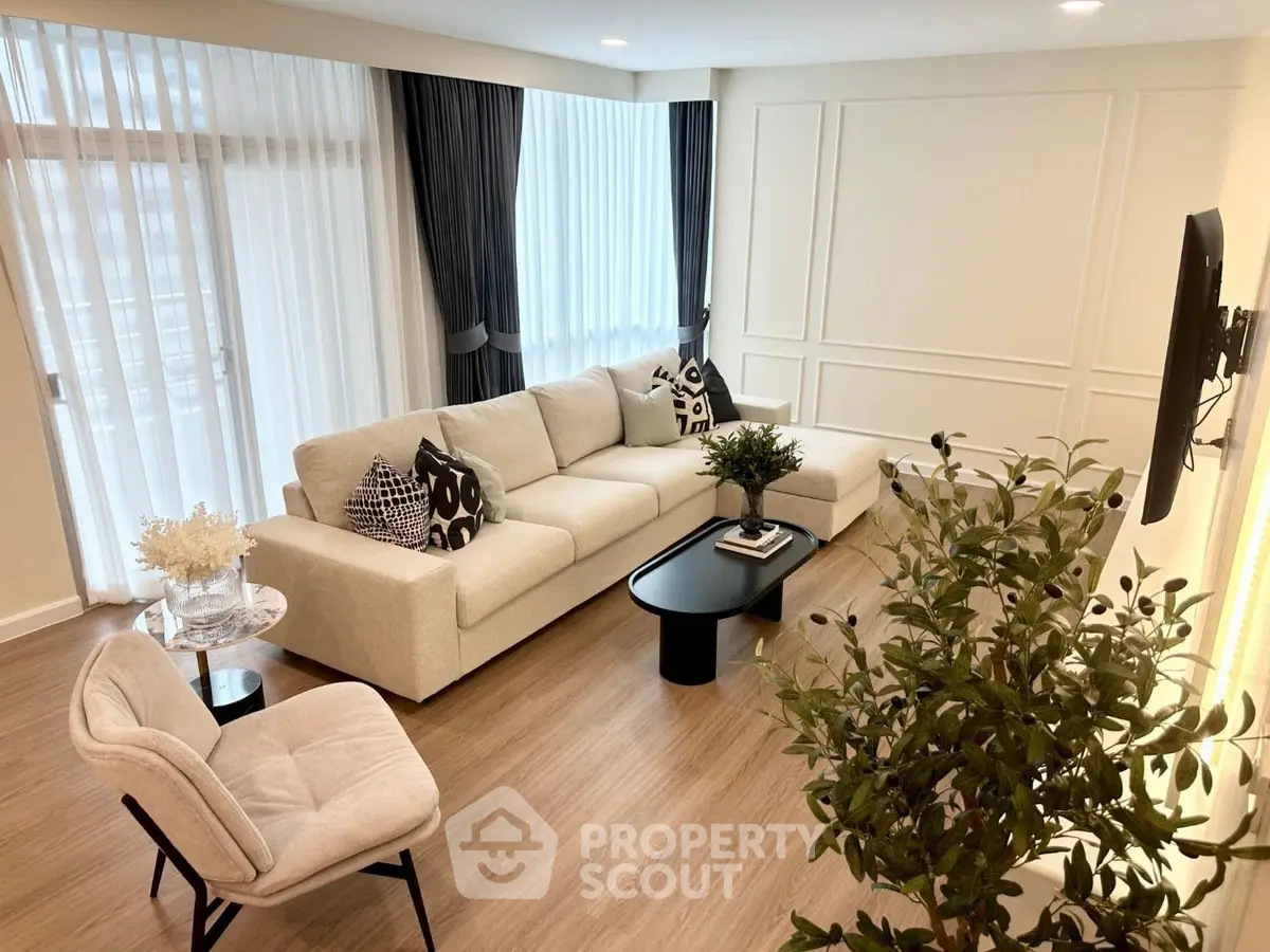 Elegant living room with modern decor, featuring a spacious beige sofa, stylish armchair, and large windows with curtains.