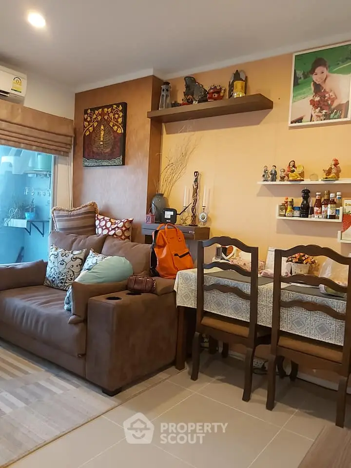 Cozy living room with brown sofa and dining area, featuring decorative shelves and warm lighting.