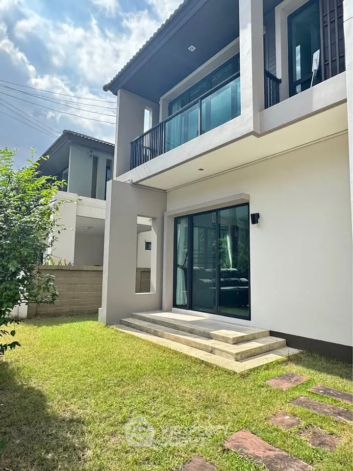 Modern two-story house with balcony and lush green lawn under a clear sky.