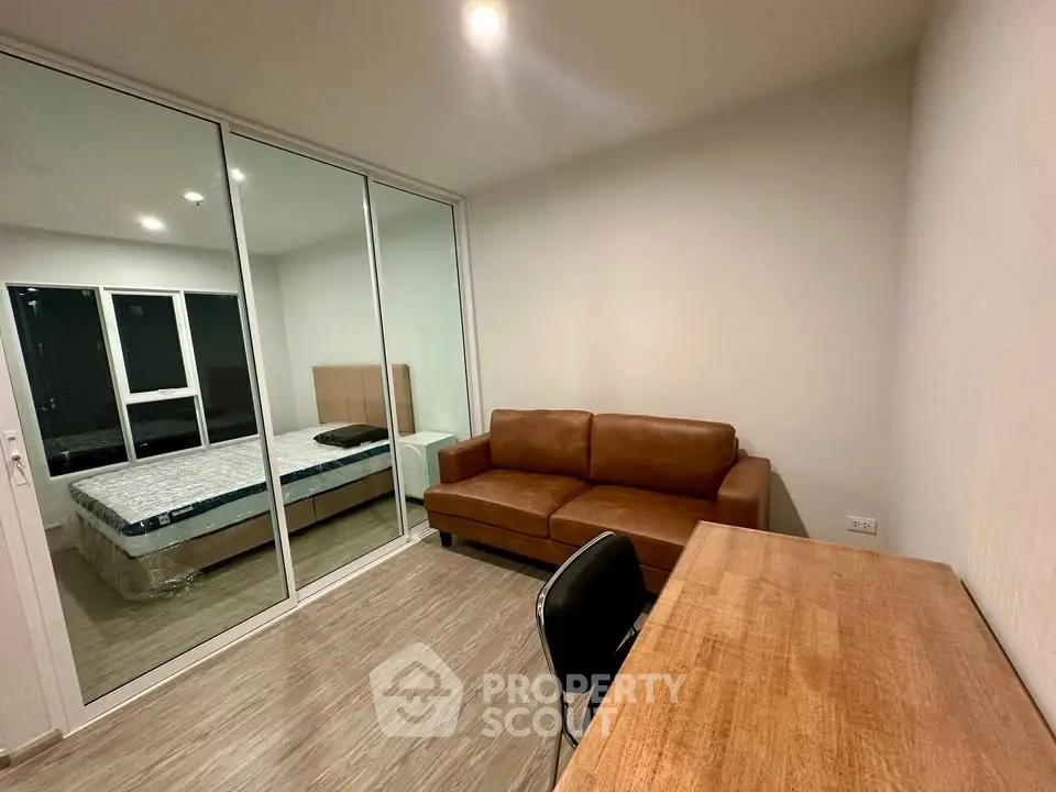 Modern apartment with cozy living room and bedroom separated by glass wall, featuring a brown leather sofa and wooden desk.