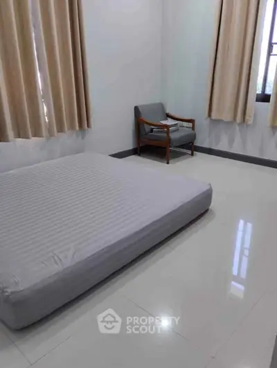 Minimalist bedroom with mattress and armchair, bright natural light through large windows.
