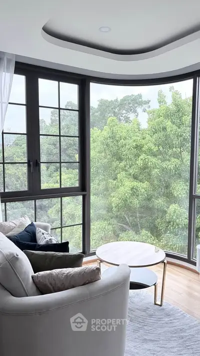 Bright living room with large windows offering lush green views, modern furniture, and cozy ambiance.