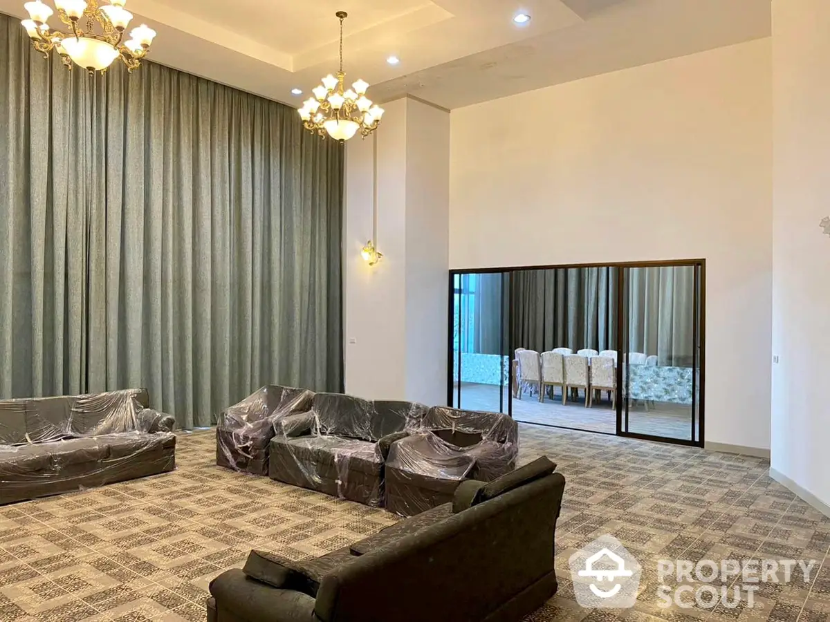 Spacious and elegant living room with large windows draped in luxurious curtains, complemented by a chic chandelier and a reflective glass partition leading to a dining area.