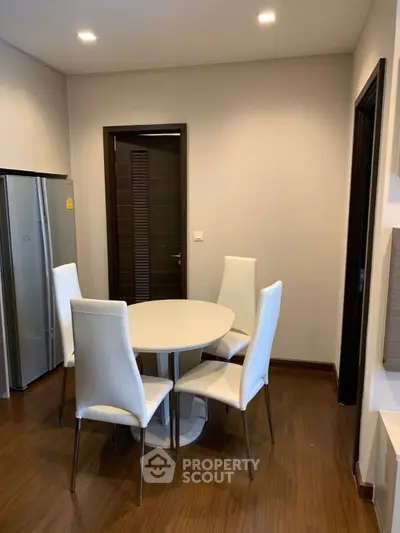 Modern dining area with sleek white chairs and round table, adjacent to a stainless steel fridge.