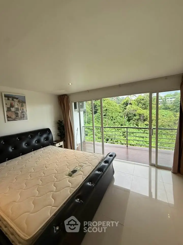 Spacious bedroom with large windows and balcony overlooking lush greenery.