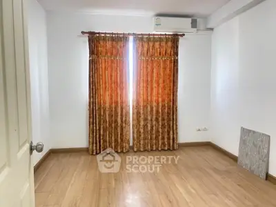 Spacious and well-lit empty room with polished wooden flooring, large windows draped with orange curtains, and a clean white ceiling with an air conditioning unit.