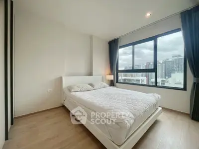 Modern bedroom with large windows offering city views, featuring a cozy white bed and wooden flooring.