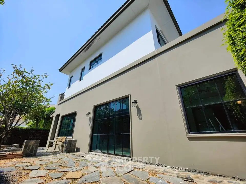 Modern two-story house with large windows and stone patio in a serene garden setting.