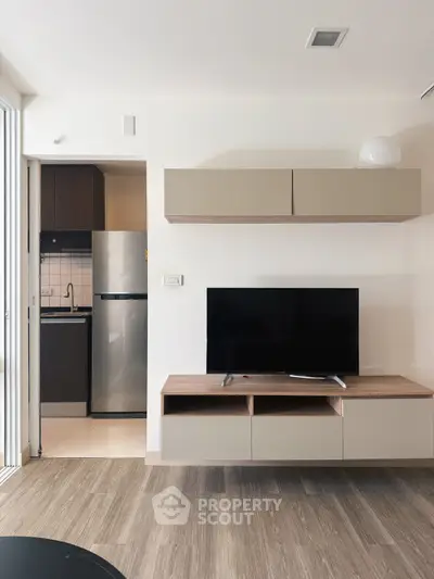 Modern living room with sleek TV unit and view into compact kitchen with stainless steel fridge.
