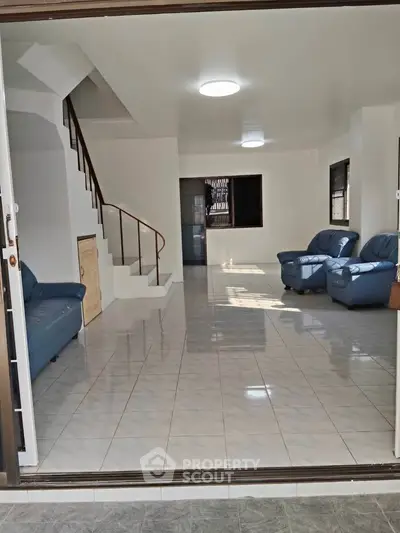 Spacious living room with tiled floors and blue leather sofas, featuring a staircase and ample natural light.