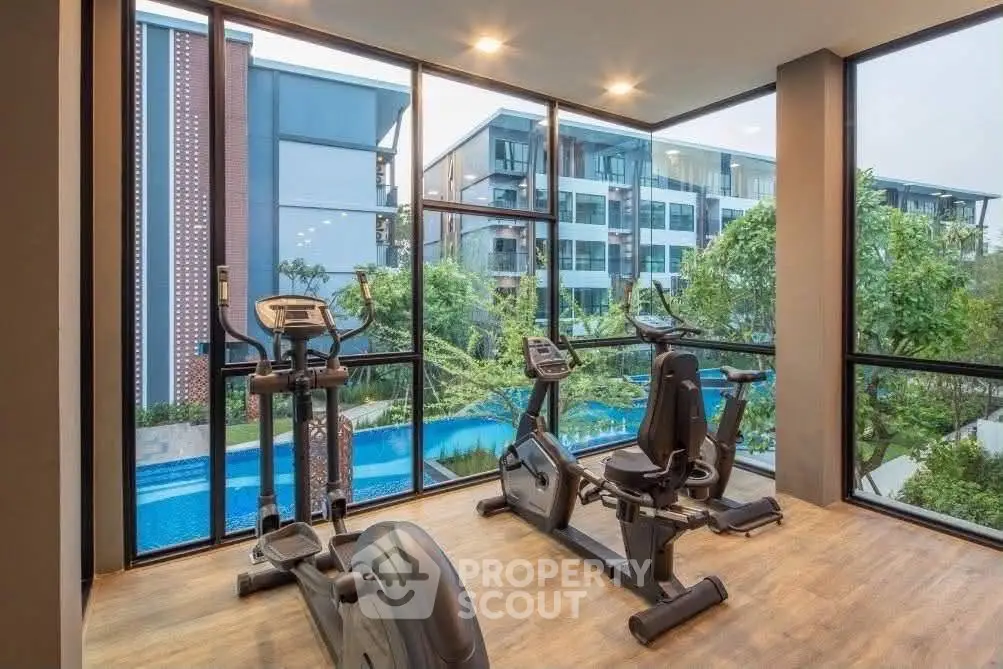 Modern gym with large windows overlooking a pool and garden in a luxury apartment complex.