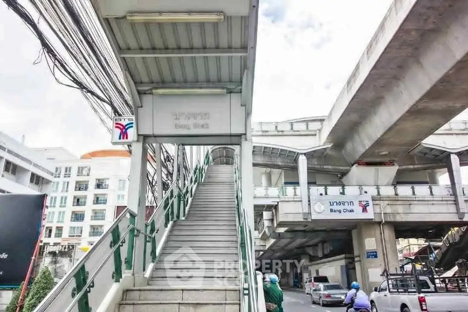 Urban transit station entrance with stairs leading to elevated platform, surrounded by city buildings and traffic.
