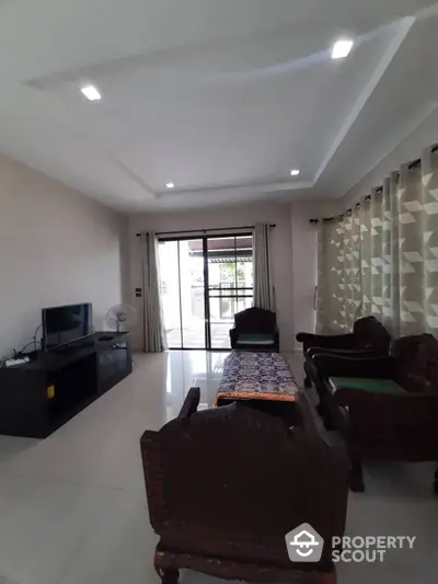 Spacious living room with modern lighting, elegant curtains, and classic wooden furniture leading to a sunlit balcony.