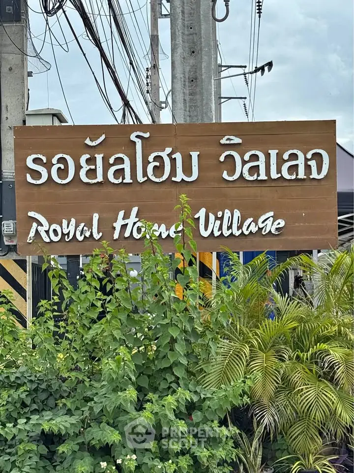 Entrance sign for Royal House Village with lush greenery and power lines in the background.