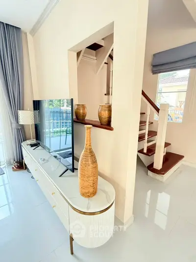 Stylish modern living room with elegant decor and staircase, featuring a sleek TV stand and large window.