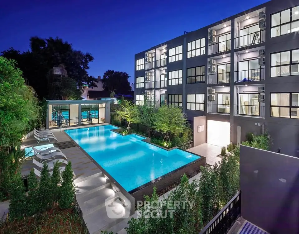 Modern apartment building with illuminated pool and lush greenery at night.