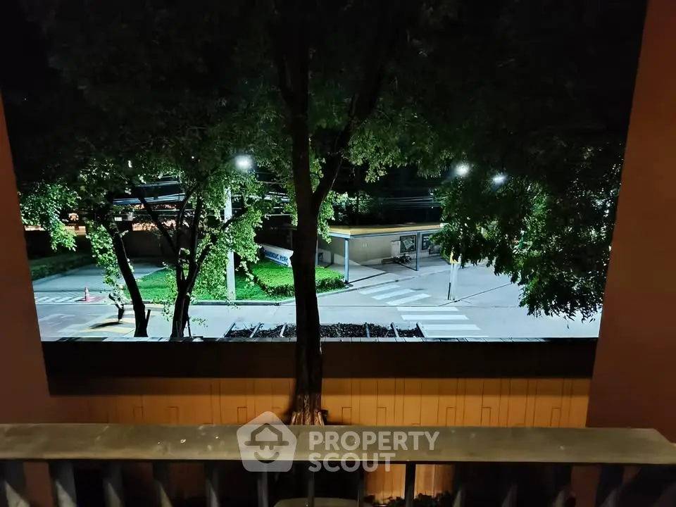 Charming nighttime view from a balcony overlooking a quiet street with lush trees and ambient lighting.