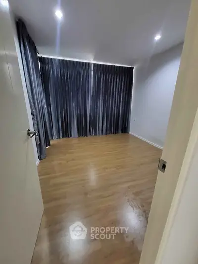 Spacious empty room with wooden flooring and large window covered by dark curtains.